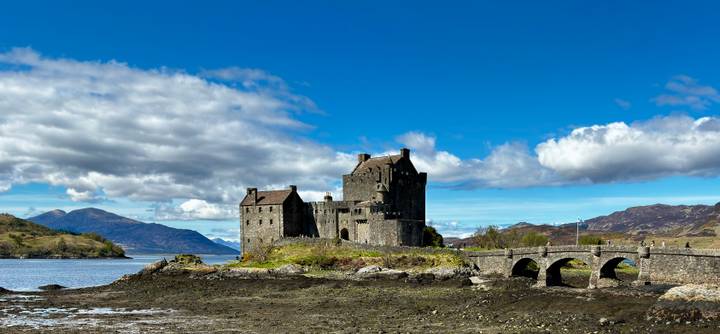 Historic Eilean Donan Castle sits on a rocky islet with surrounding mountains and bridge under blue skies.