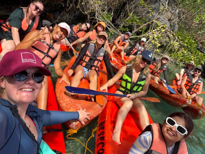 Smiling travellers in life jackets sit in bright orange kayaks waiting to paddle crystal clear water.