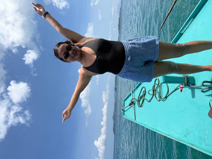 Woman stands joyfully on the bow of a turquoise boat surrounded by calm tropical sea and blue sky.