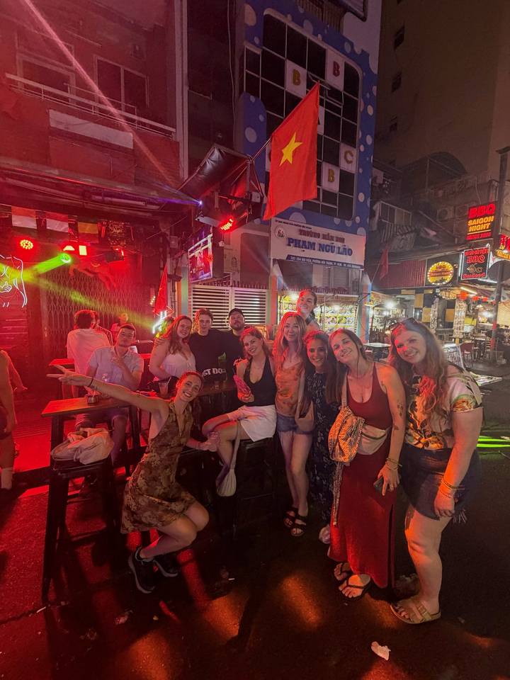 A lively group of backpackers poses inside a neon-lit bar street at night.