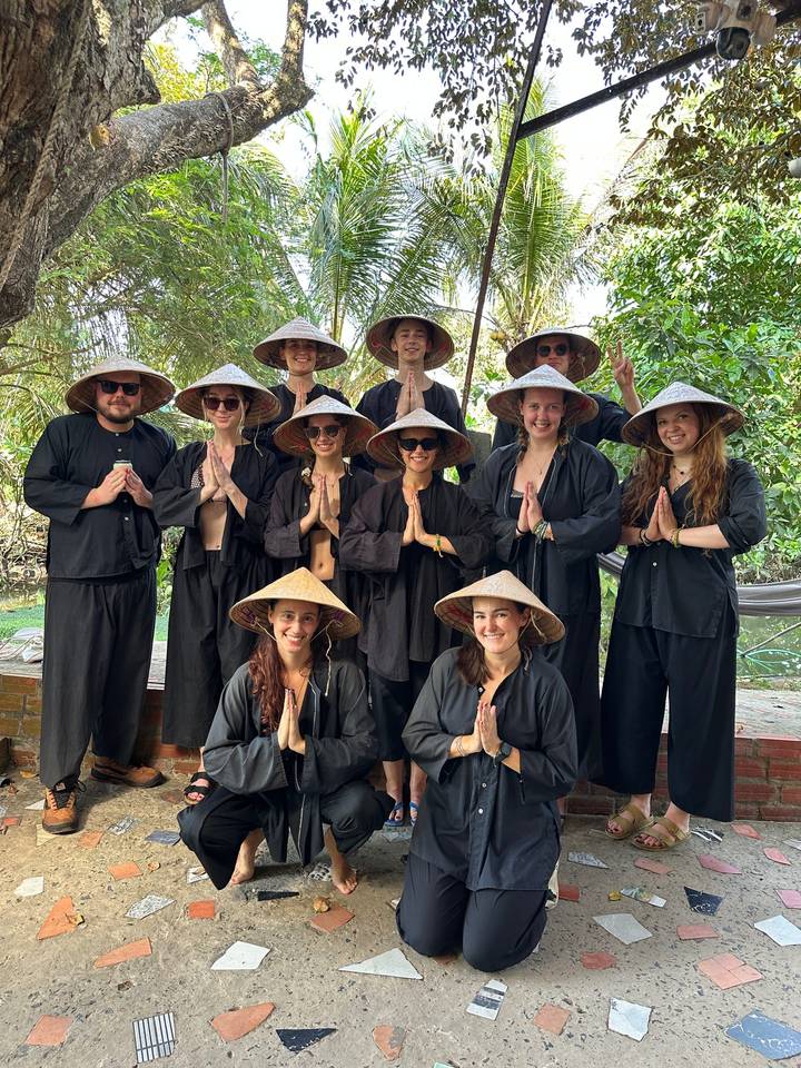 Travellers dressed in traditional black clothing and conical hats pose with palms together in a tropical setting.