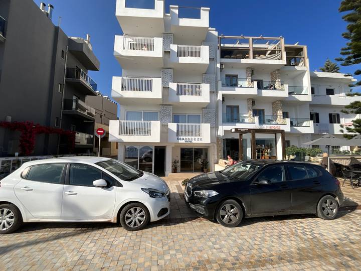 Facade of a small seaside apartment hotel with parked cars on a sunny street in Crete.