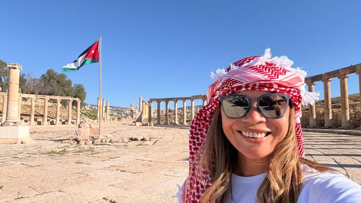 Selfie of a smiling woman wearing a red-and-white keffiyeh with Jerash’s oval colonnade and Jordan flag behind her.