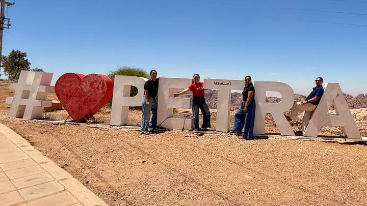 Travellers stand beside a large #❤️PETRA sign under clear blue skies.