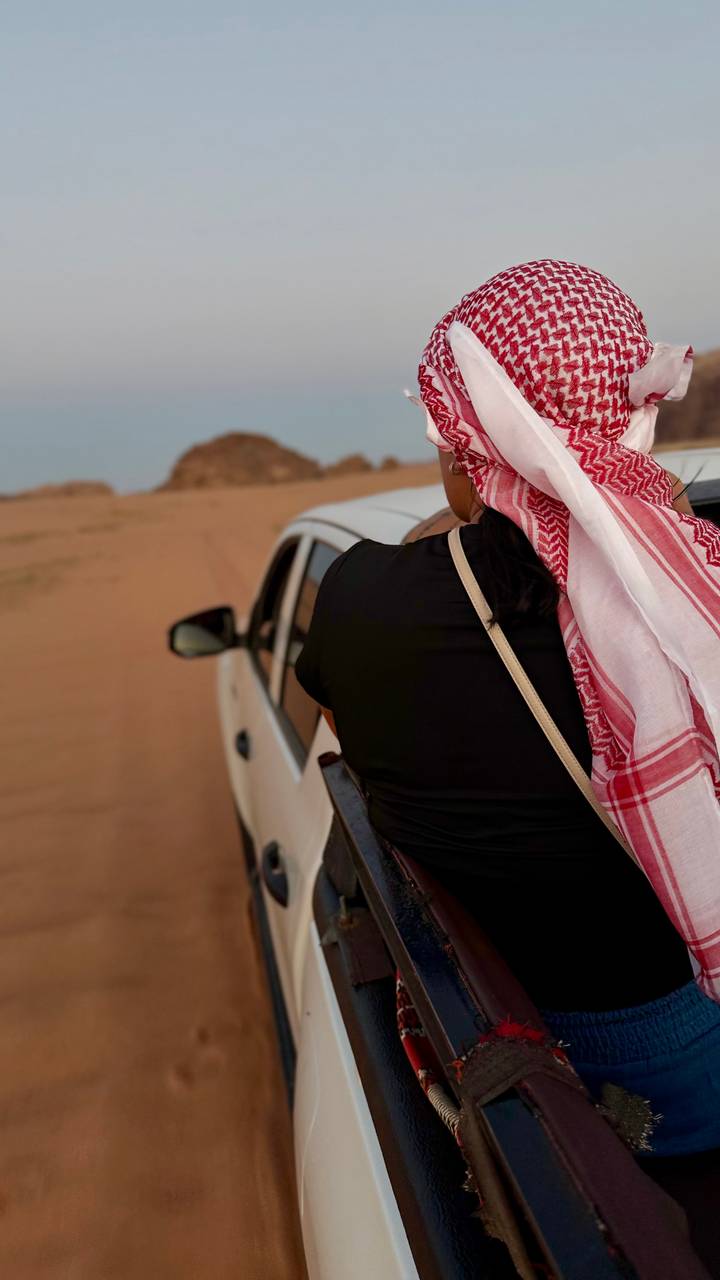 Passenger wearing a red-and-white scarf leans out of a white car driving across desert sands.
