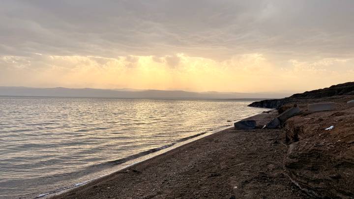 Calm Dead Sea shoreline at dusk with golden rays breaking through clouds across the water.