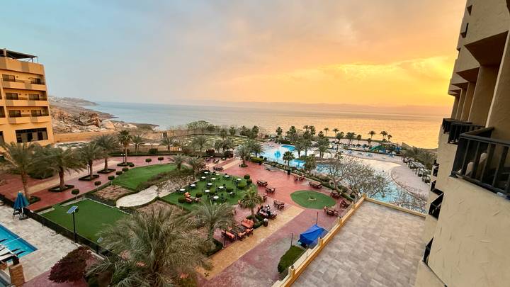Resort gardens, pools and palm trees overlook the Dead Sea under a colourful sunset sky.