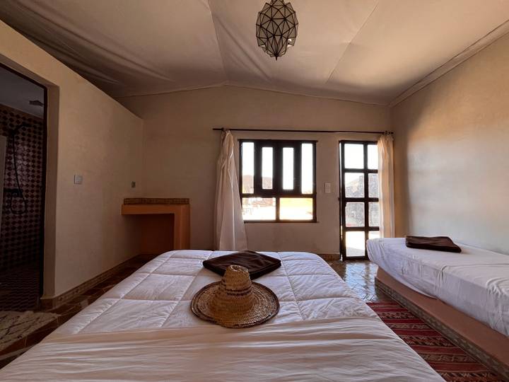 Simple desert lodge room with twin beds, woven hat and sunlight filtering through the window.