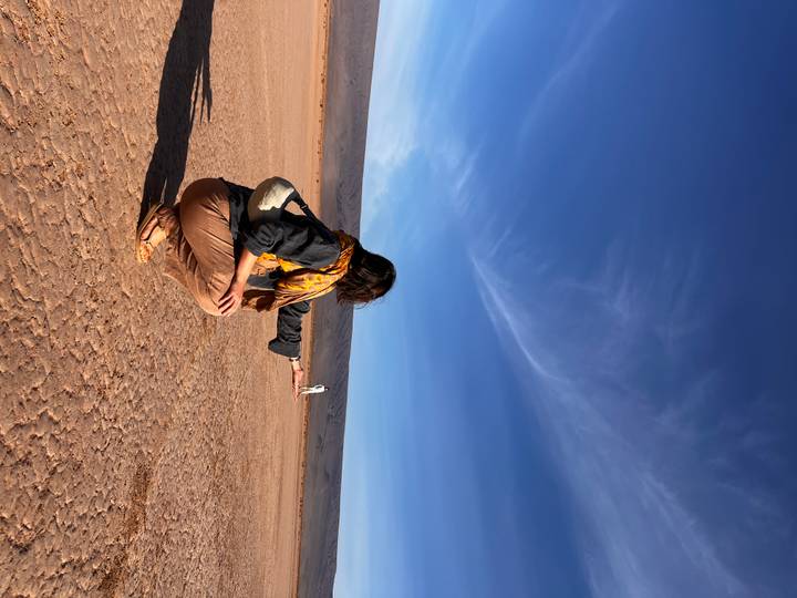 Traveller squats on cracked desert ground creating a forced-perspective pose with a distant friend under vivid blue sky.