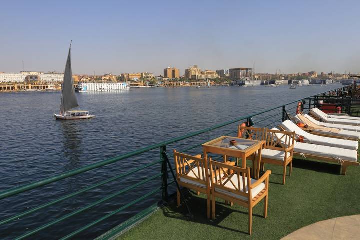 Sun loungers and tables line the deck of a Nile cruise boat overlooking calm river waters and a passing felucca sailboat.