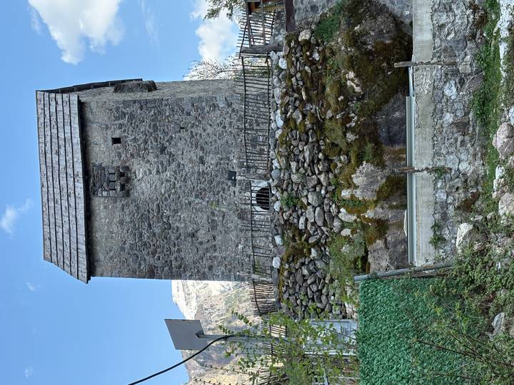 A stone defensive tower house rises above a moss-covered wall in an alpine village.