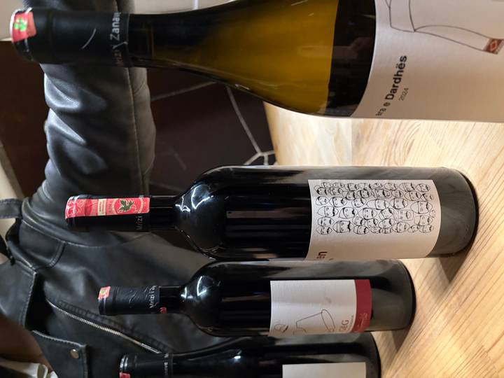 Bottles of Albanian wine stand on a wooden bar while an arm in a leather jacket rests behind them.