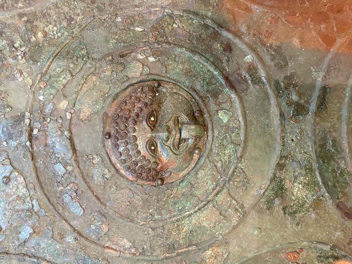 Close-up of an ancient bronze shield or artefact featuring a gorgon face with tongue protruding.