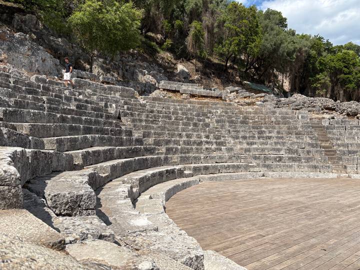 Ancient stone amphitheatre tiers curving around a wooden stage with one visitor walking along the top row