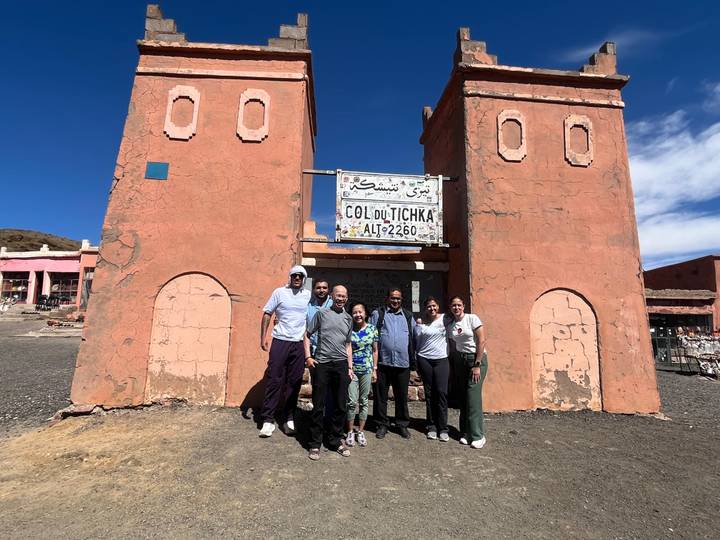 Travel group poses by the Col du Tichka pass sign amid Atlas mountain scenery.