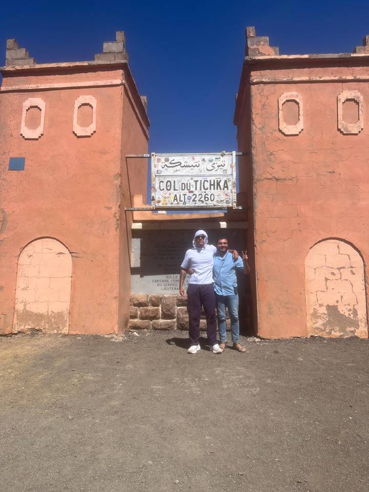 Two men pose in front of a hazy Col du Tichka altitude sign in bright desert light.