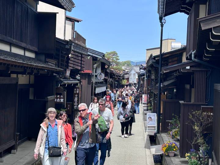 Busy traditional street in Takayama with wooden merchant houses and a crowd of tourists walking under blue skies.