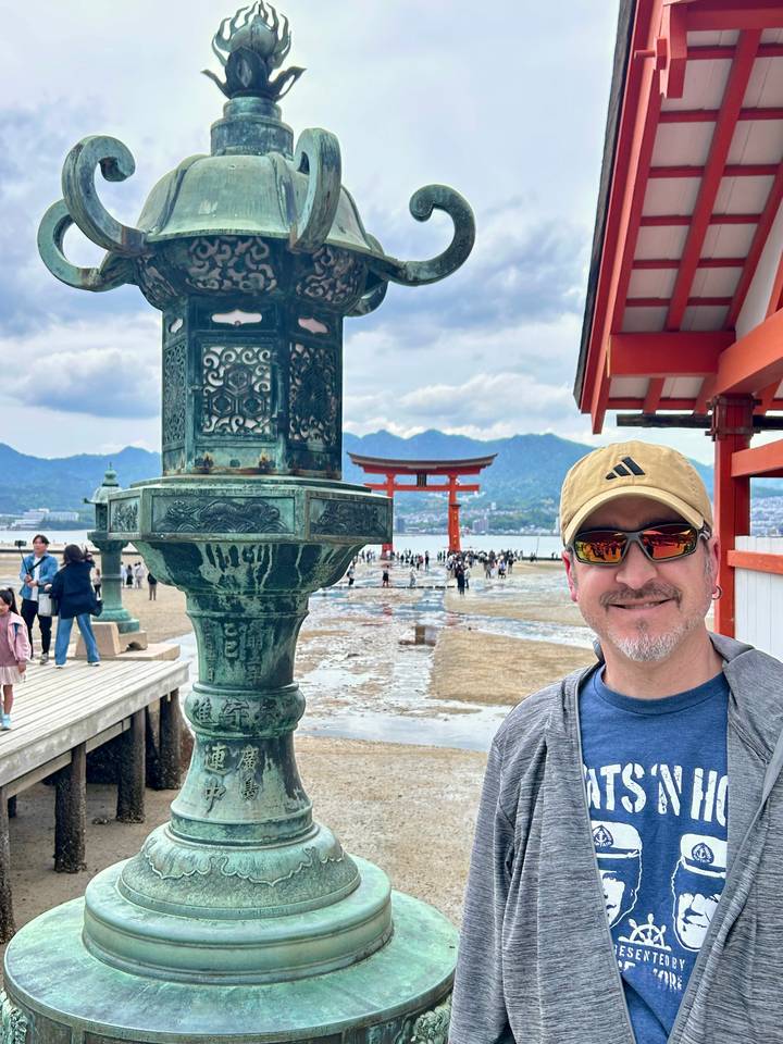 Smiling tourist in front of bronze lantern with the famous floating Torii gate of Miyajima Island and distant mountains.