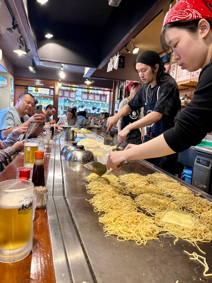 Chefs prepare heaps of noodles on a large teppan grill while diners sit at the counter in a bustling Japanese eatery.