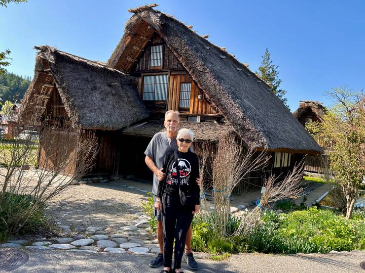 Older couple poses in front of a traditional gassho-zukuri thatched farmhouse in Shirakawa-go on a sunny morning.