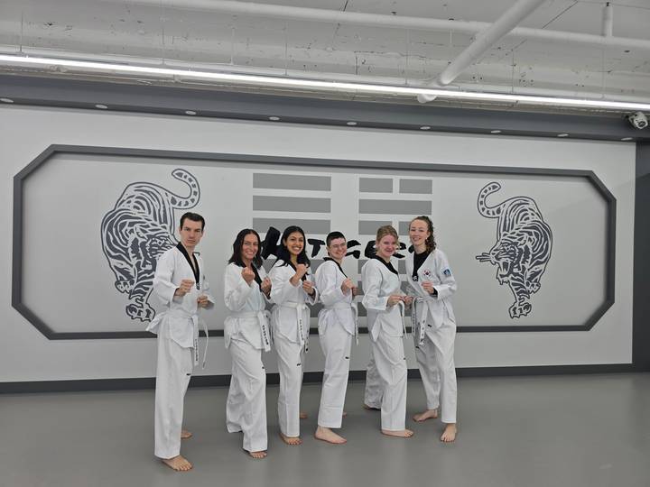 Travelers dressed in white taekwondo uniforms posing proudly in a modern dojang decorated with tiger murals.