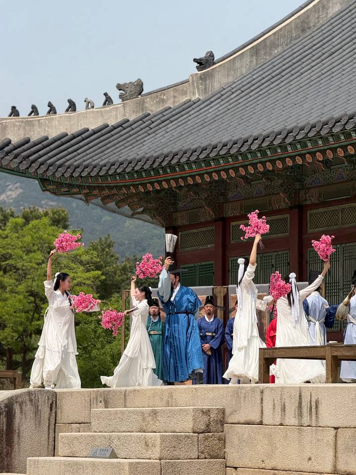 Traditional Korean dance performance with women in flowing hanbok waving pink blossoms in a palace courtyard.