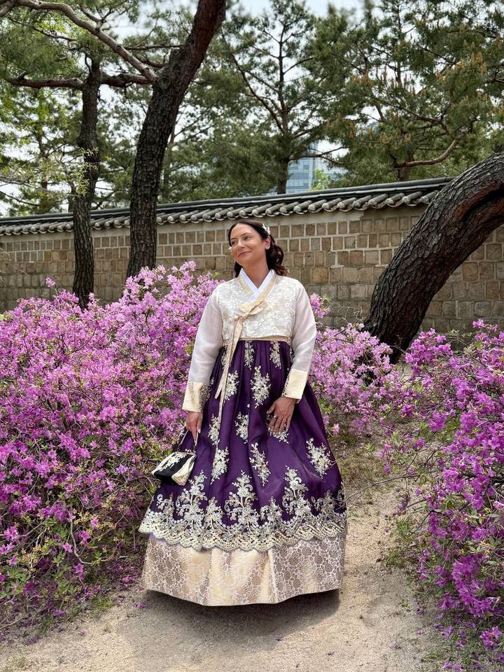 Woman wearing traditional hanbok poses among blooming pink azaleas beside a stone wall.