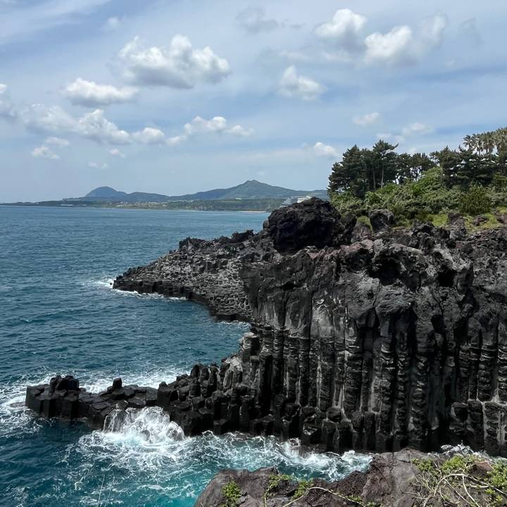 Dramatic basalt column cliffs meeting a deep blue ocean with distant mountains under a partly cloudy sky.
