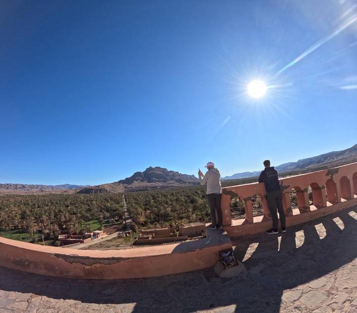 Two travelers on a terrace taking photos over palm oasis and distant mountains.