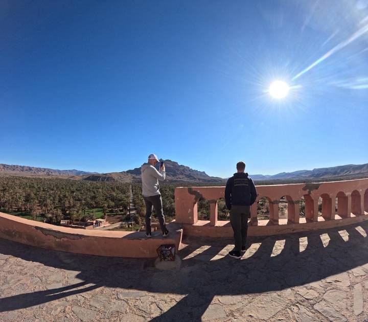 Two travelers photographing desert oasis panorama from pink parapet.