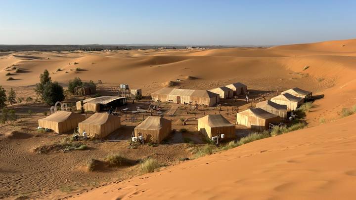A cluster of beige tented huts forms a desert camp amid sweeping orange dunes under a clear sky.