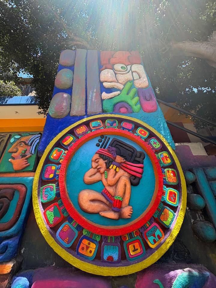 Vivid Mayan-style relief mural with carved calendar ring and reclining figure on a bright column.