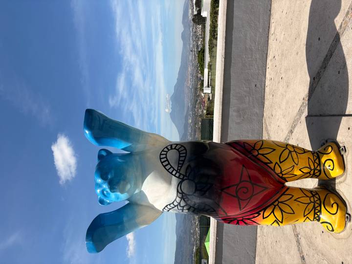 Colorful painted bear statue with arms raised on a rooftop terrace overlooking a valley city and a distant conical volcano.