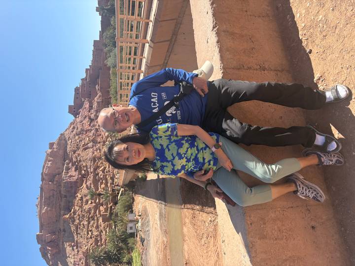 Couple posing on a bridge with the clay-colored kasbah structures of Aït Benhaddou rising on a rocky hill behind them.