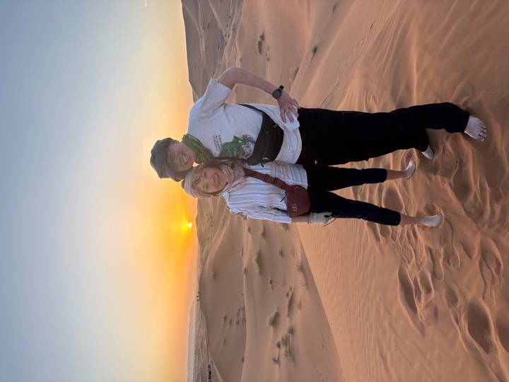 Couple standing barefoot on sand dunes at sunset with the sun dipping below the horizon in the Sahara Desert.