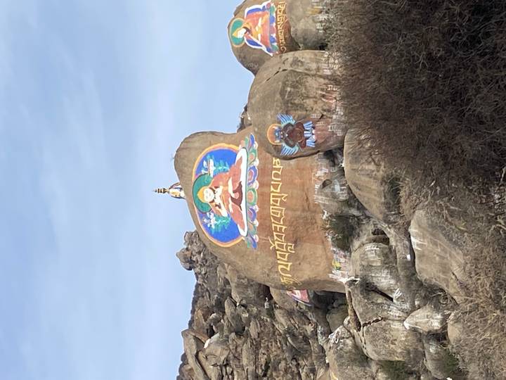 Colorful Buddhist deity mural painted on a large boulder set into a rocky hillside.