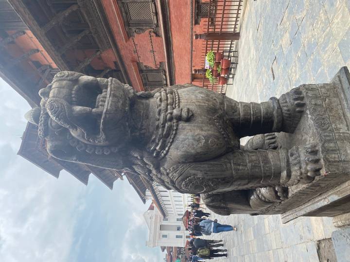 Stone guardian lion statue in Kathmandu Durbar Square with tourists exploring historic brick palaces.