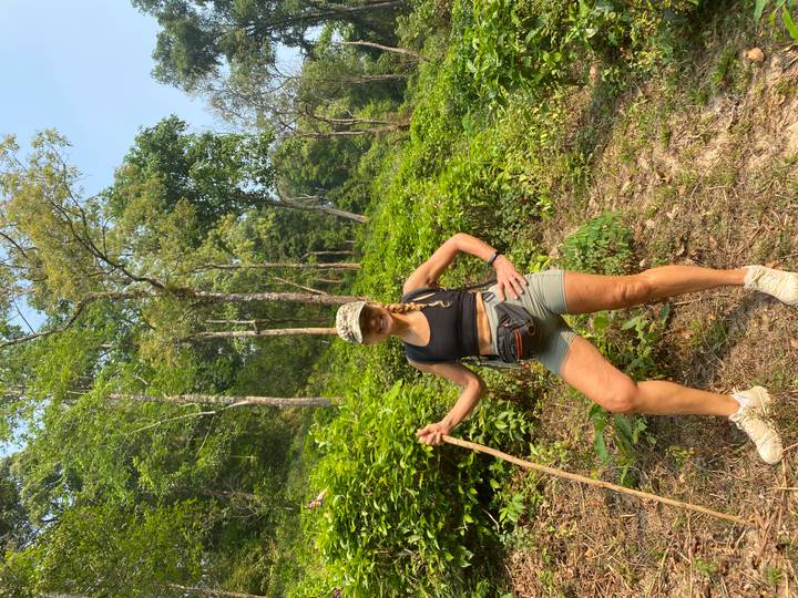 Smiling trekker posing with walking stick in lush green forest terrain.
