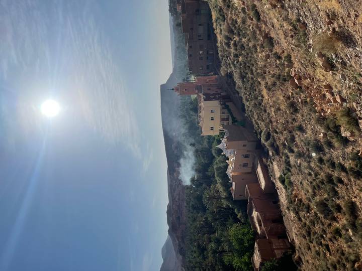 Sunlit desert village with mosque minaret, palm groves and smoke rising in a rocky valley.