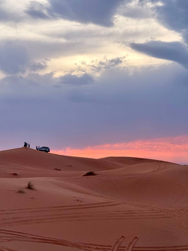 Silhouettes of people and a 4x4 vehicle atop a desert dune against a vivid pink sunset sky.
