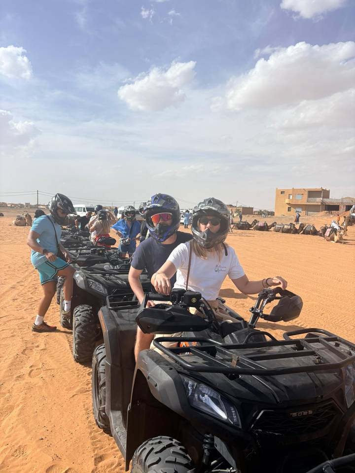 Travelers wearing helmets sit on quad bikes lined up on desert sand ready for a ride.