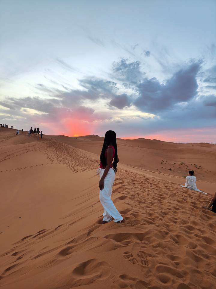 A woman gazes at a vivid sunset over rolling sand dunes as camel riders crest a ridge.