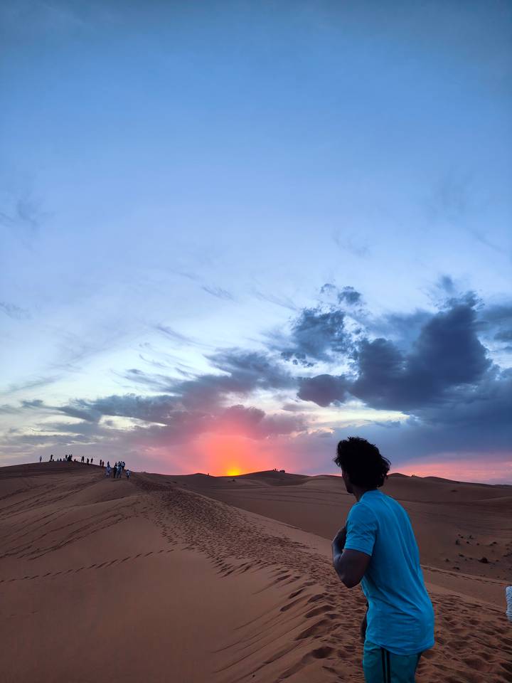 Silhouettes of travelers line a dune against a dramatic sky as the sun sinks below the horizon.