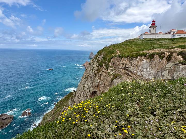 Dramatic ocean cliffs capped by a lighthouse at Cabo da Roca with wildflowers in the foreground.