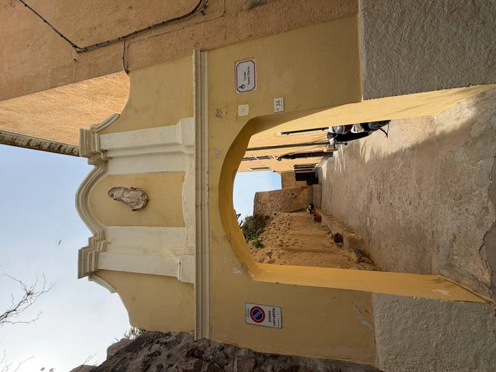 Yellow stucco archway leading into a narrow lane with stone walls and potted plants.