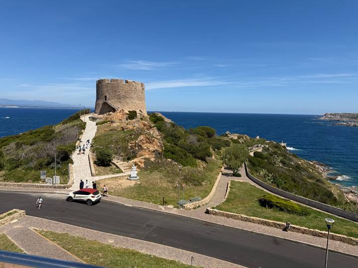 Stone coastal watchtower on a rocky promontory overlooking deep blue Mediterranean waters with visitors walking the path.