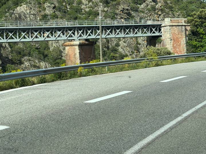 Close-up of a highway curve with metal guardrail and railway bridge in the background.