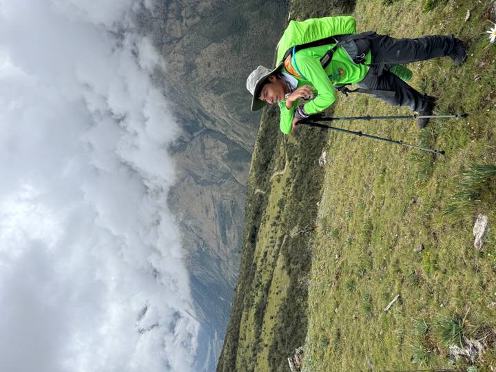 Hiker in green jacket resting on trekking poles while gazing at misty mountain valley.