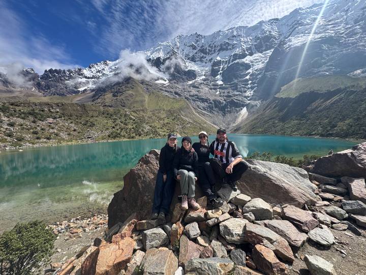 Four travelers sitting on rocks by a vibrant emerald lake with snowy peaks above.