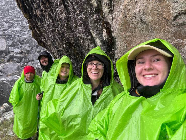 Smiling hikers wearing bright green ponchos sheltering by a rock wall in rainy weather.
